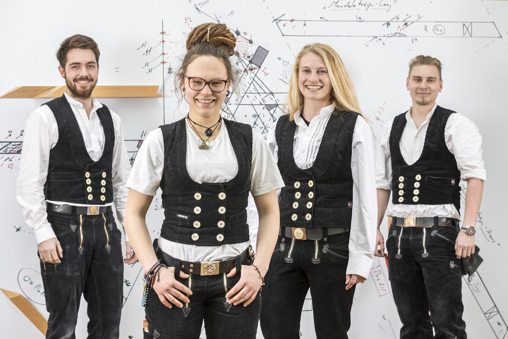 Vier Auszubildende von Holzbau Trunzer GmbH in traditioneller Zunftkleidung vor einer Tafel mit technischen Zeichnungen