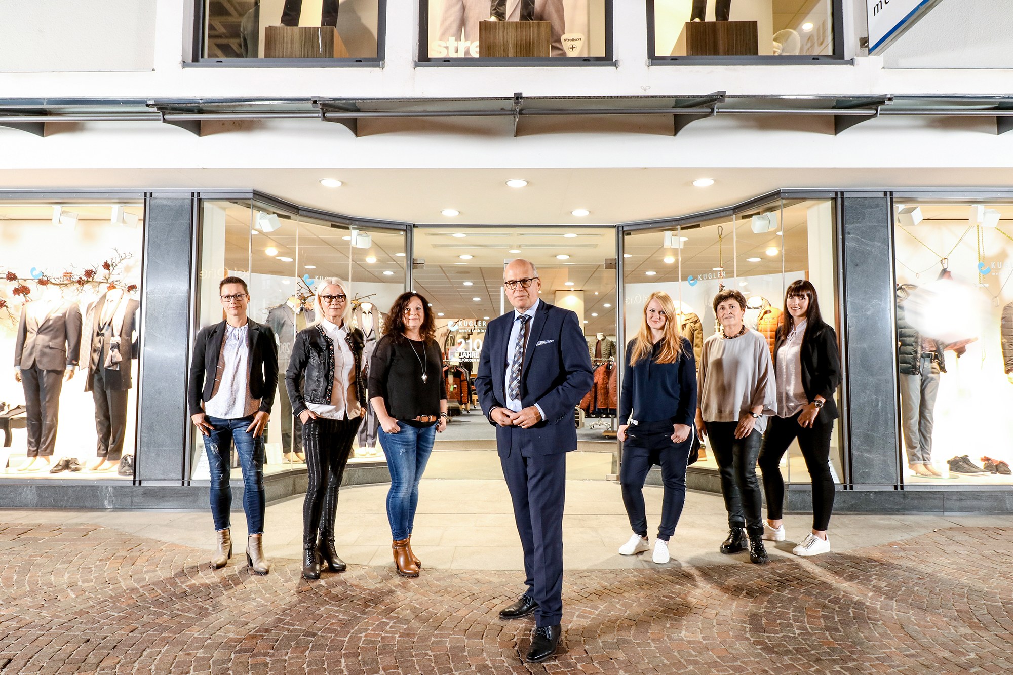 Team von Kugler Men's Fashion vor dem beleuchteten Schaufenster des Modehauses