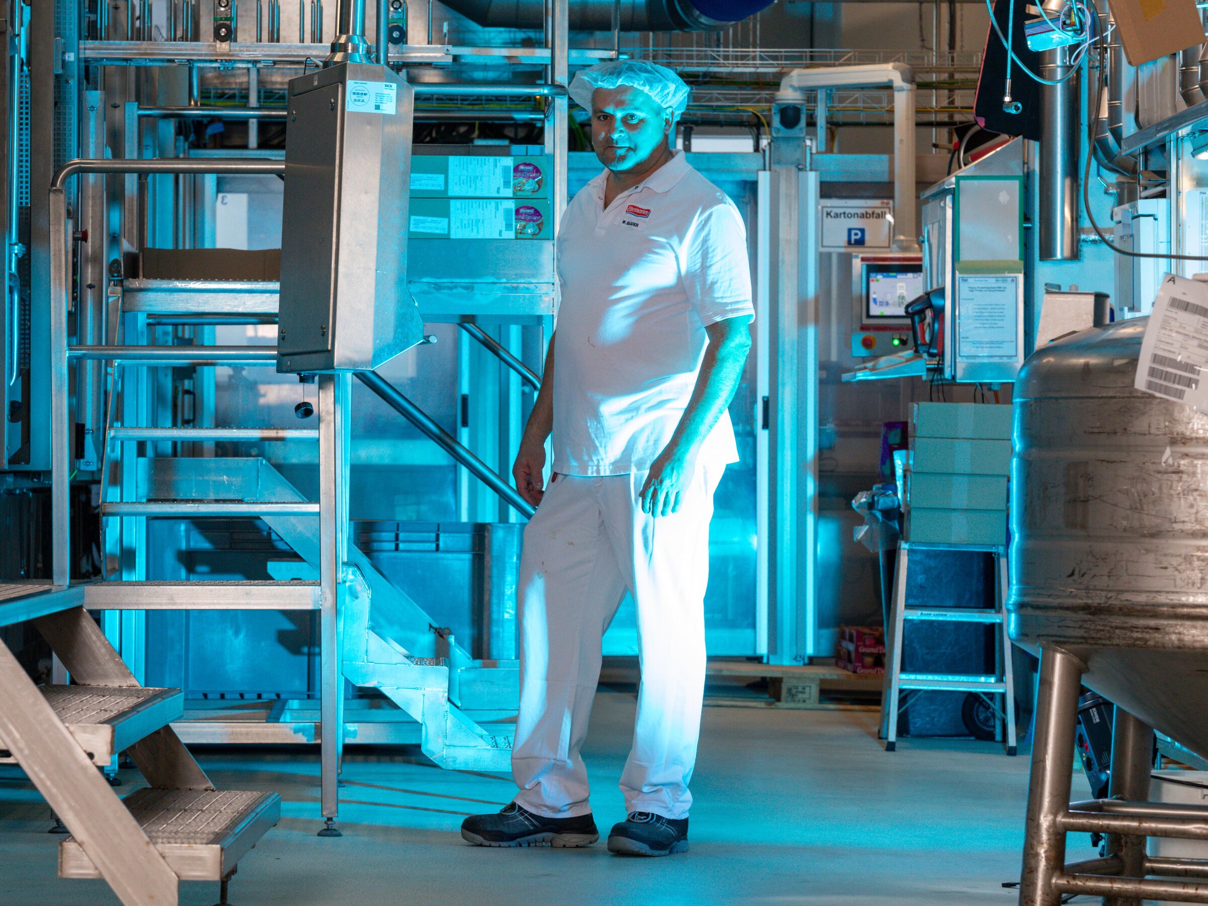 Mitarbeiter in Produktionshalle blickt konzentriert zur Seite, blau beleuchtete Industrieumgebung mit Edelstahlanlagen